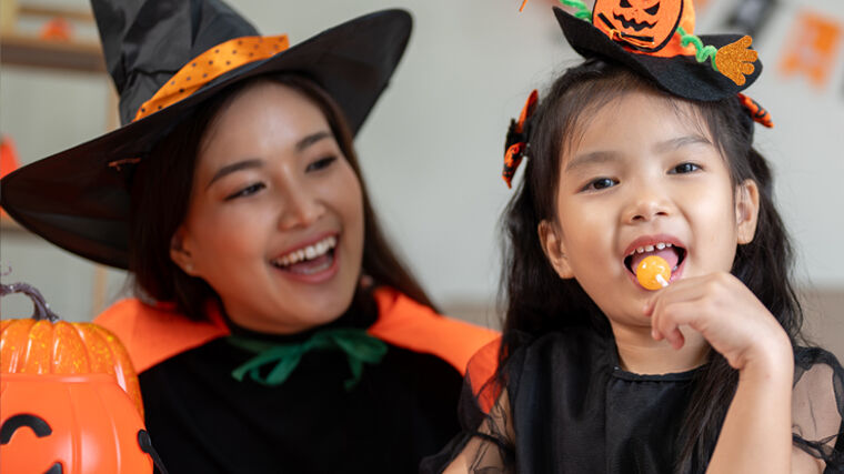 Person in a black-and-orange witch costume sits beside a child in a Halloween outfit who is holding and eating an orange lollipop.