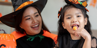Person in a black-and-orange witch costume sits beside a child in a Halloween outfit who is holding and eating an orange lollipop.