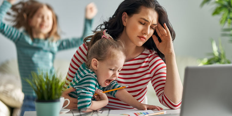 Stressed out mother sits at a table, young daughter sitting on her lap, child in background