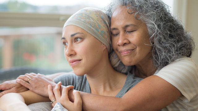 Older woman hugs a younger woman with cancer, wearing a head scarf.